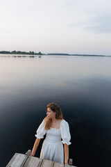 Fototapeta premium A woman in a white dress stands in the water near a wooden pier, holding onto the edge. The evening sky reflects in the calm lake. In the distance, a boat with people adds to the sense of solitude