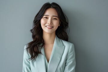 Fototapeta premium A Young Asian businesswoman in a pastel green suit poses confidently against a soft grey-green backdrop, embodying leadership and fashionable office attire, ideal for corporate use.
