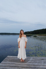 A young woman in a white dress stands barefoot on a wooden pier by a lake, surrounded by lilies and forest. The evening sky creates a peaceful atmosphere. She gazes thoughtfully into the distance.