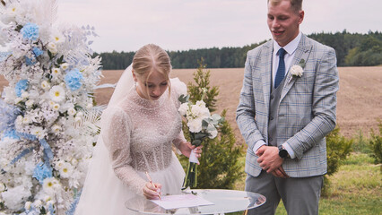 Bride signing marriage certificate during outdoor wedding ceremony with groom looking on, celebrating their love and commitment