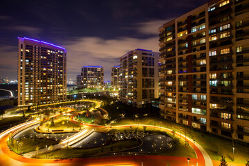 Modern high-rise residential buildings surrounding a beautifully illuminated urban park with winding paths, landscaped gardens, and decorative water features under a night sky in a vibrant city