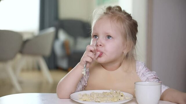 Cute little baby blonde hair girl having breakfast, eating corn flakes, drinking fresh milk, licks thumb in the kitchen in the morning. Healthy eating habits. Children nutrition footage