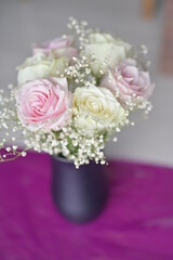 Delicate bouquet of soft pink and creamy white roses adorned with tiny baby's breath flowers in a sleek black vase set against a vibrant purple background creating a beautiful and elegant display 