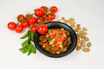 Delicious Mediterranean Fava Bean Stew with Fresh Tomato Salsa, Chickpeas, and Herbs in a Black Bowl, Surrounded by Ripe Cherry Tomatoes on the Vine, Fresh Mint Leaves, and Raw Fava Beans