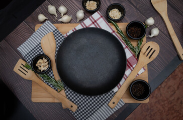 Aesthetic rustic kitchen setup with a black empty plate, wooden cooking utensils, fresh rosemary, garlic, nuts, and spices on a dark wooden table with checkered and striped kitchen towels.