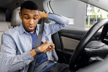 Got stuck in traffic jam. Stressed black man sitting on drivers seat, looking at watch on his wrist and touching head, being late