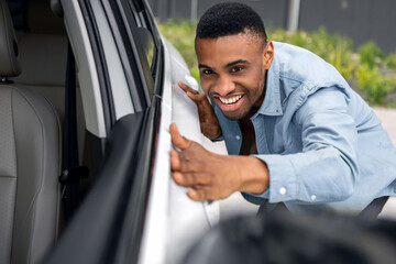 Happy young black man buying new car, checking automobile surface and smiling, guy looking at...