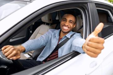 Happy African American man driving luxury car, showing thumb up and smiling, sitting inside...