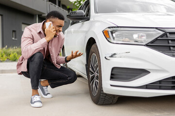 Young angry black man sitting near scratched car and calling car service, trying to solve problem...