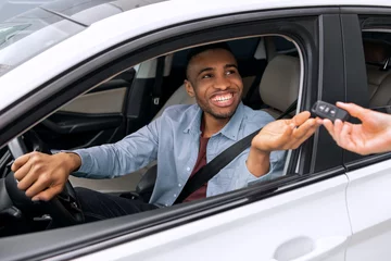 Happy African American man taking key from seller buying auto in dealership center, sitting inside and smiling. Guy doing test-drive before buying new automobile