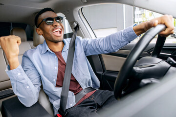 Overjoyed African American guy in sunglasses dancing in car, listening music and singing while...