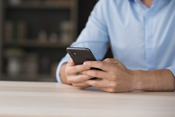 Working in phone app. Close up cropped shot young man sit at desk hold mobile telephone in hands communicate via internet manage tasks make call stay connected type message text on screen. Copy space