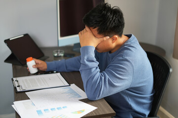 A young asian manager is taking medicine while sitting in front of laptop at office, looking for treatment against coronavirus, cold or flu and has been suffering headache due to heavy work load. 