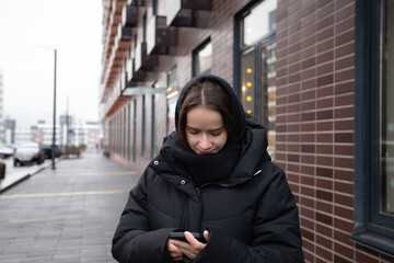 Young Woman in Black Winter Jacket Using Smartphone on City Street in Cold Weather