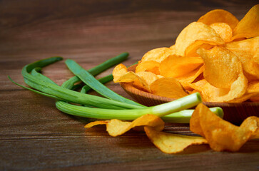 A plate of potato chips sits on a wooden table with a bunch of green onions. The chips are scattered all over the table and the onions are stacked in the background.