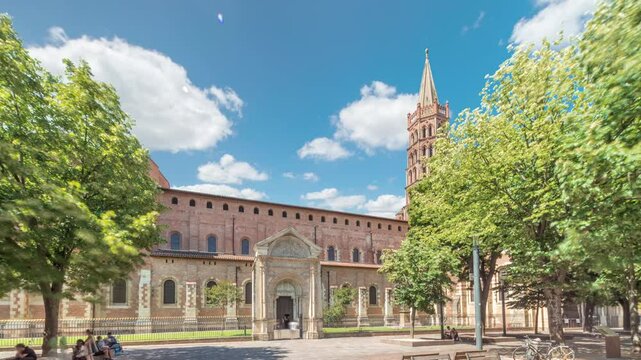 The cobbled parvis of the Basilica of Saint Sernin in Toulouse, France, timelapse hyperlapse. Largest romanesque church in Europe with brick bell tower, arched doors, green trees and blue cloudy sky