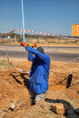 Single African road construction worker with a pickaxe digging a trench ditch on the side of the...