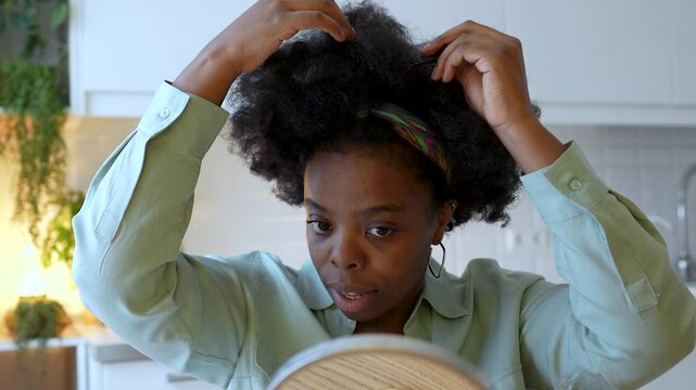 Focused African American woman styling natural afro hair with wide-tooth comb and mirror at home. Self-care, individuality, beauty routine in kitchen with houseplants, embracing curls care, self-love.