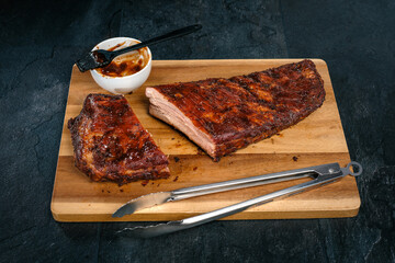 St. Louis Cut Ribs auf Holzbrett, Holzteller mit Grillzange, Sch&uuml;ssel mit Barbecue Sauce und Pinsel auf schwarzem Schiefer