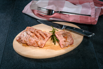 Angeschnittenes Flanksteak vom Schwein auf ovalem Holzteller mit Messer, Gabel, Stoffserviette und...