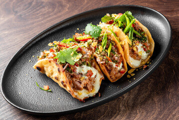 A close-up of three delicious tacos filled with savory meat, fresh vegetables, and garnished with herbs, served on a sleek black plate against a wooden background