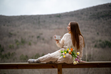 Young woman enjoys tea on a deck with mountain view, wearing a knit sweater and surrounded by blooming tulips in early spring