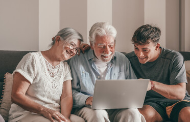 Cheerful Multi generation family of grandparents and young grandson share social media on laptop sitting together on sofa at home. Old couple learning use of computer and technology