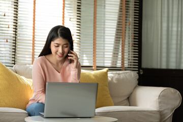 Asian young woman sitting on cozy sofa, smiling while talking on phone and working on laptop in...