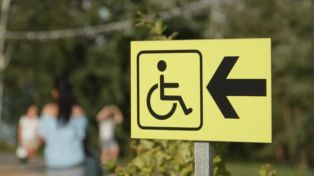 Sign for direction of path for people with disabilities in city park. Blurred silhouettes of unrecognizable people against bright yellow information sign with arrow for disabled.