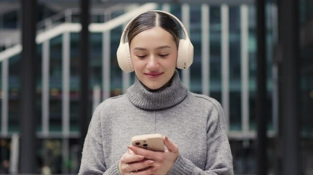 Chill mode in the fast lane. Headphones on, sweater game strong, a woman scrolls her phone outside glass-fronted buildings, merging comfort with metropolitan cool