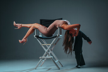 Young woman gymnast in elegant dress stretching on high chair on gray wall background
