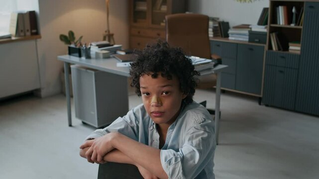 Medium portrait of 12 year-old curly-haired Black troublemaker schoolboy posing on chair, child with torn shirt and bandaged nose after fight, waiting in psychologists office, copy space