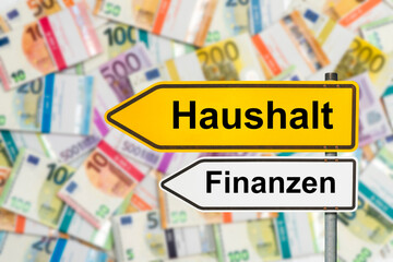 Ein Wegweiser mit der Aufschrift Haushalt und Finanzen steht vor vielen Euro Geldbündel