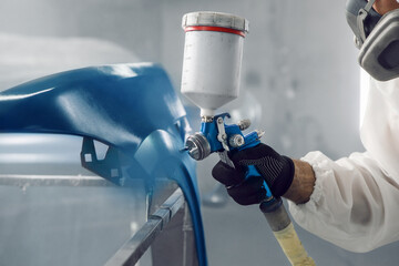 Automobile repairman painter in protective workwear and respirator painting part of car in paint...