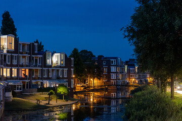 Fototapeta premium A serene canal scene at dusk, with modern brick buildings illuminated by warm lights. Reflections shimmer on the water, while trees and a narrow walkway add natural charm to the urban setting.