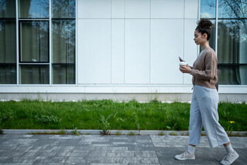 Young woman walking on sidewalk, using smartphone and holding coffee