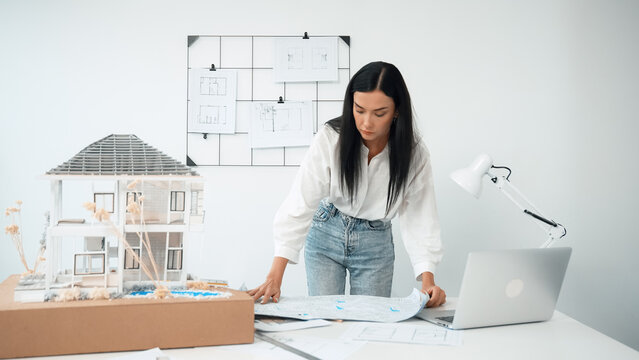 Young beautiful caucasian architect puts blueprint on the table and compare with house model while inspect hos model construction carefully at modern office with blueprint striped behind. Immaculate.