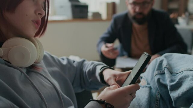 Side shot of rebellious adolescent Caucasian girl with red hair and headset on neck defiantly ignoring psychologist and rolling eyes, staring at phone and showing lack of interest in therapy session
