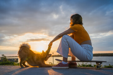 Young woman sitting on a skateboard, playing joyfully with her dog by a river during sunset,...