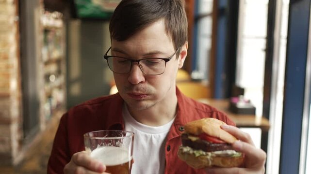 Young man savoring burger and beer at restaurant, experiencing casual dining moment with evident enjoyment and relaxation while consuming flavorful meal