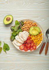 Healthy bowl with grilled chicken breast, avocado, chickpeas, corn, tomatoes and mixed greens, top view on a white wooden table from above, a balanced and nutritious meal