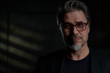 Fototapeta premium Business portrait - businessman in black jacket standing against dark gray wall. Confident middle aged, mid adult man in his 50s wearing glasses, smiling. Copy space.