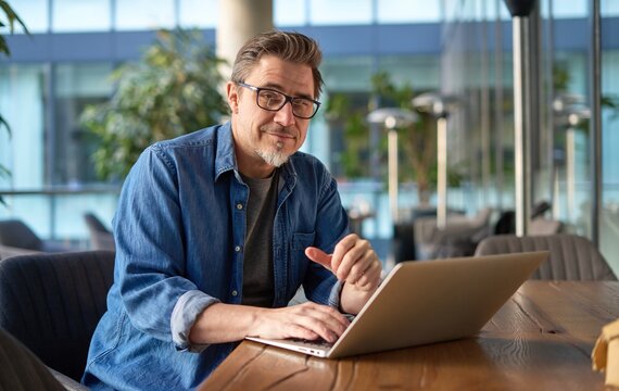 Happy middle age businessman using laptop computer in office. Older casual man working online on terrace of cafe, smiling.