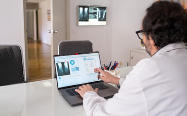 a doctor wearing a white coat types on his laptop. There is a generic health care software with x rays on the screen. In the background a viewing box with some x-rays and the corridor