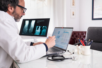 A middle-aged doctor wearing a white coat studies a case using generic anonymous healthcare software. In the background, there is another screen with some X-rays and a corridor