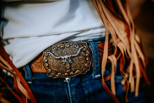 Close-up of an ornate Western-style belt buckle featuring a longhorn skull, worn with denim jeans and fringed suede vest, evoking cowboy and rodeo aesthetics..