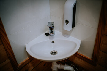 Corner bathroom sink with chrome faucet, soap dispenser, and exposed plumbing, set against tiled and wood-paneled walls in a compact washroom setup..