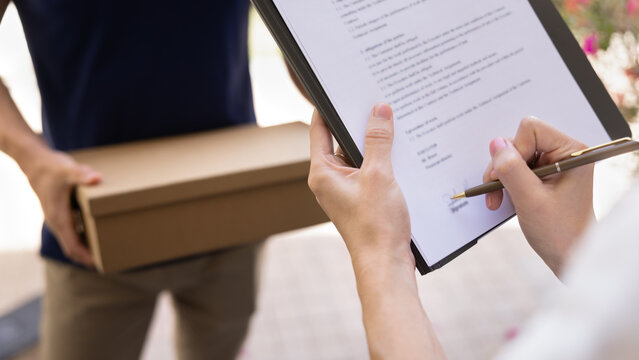 Trust and professionalism in e-commerce logistics. Close up cropped view of unknown male courier holds package box while female recipient signs confirmation form, ensuring secure and safe transaction