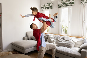 Strong father and son dressed in red capes playfully engaged in imaginative superhero game at home....