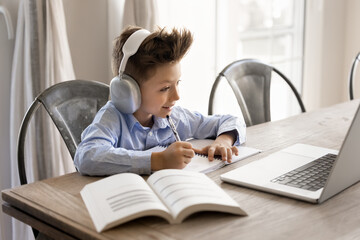 School age boy sit at table wearing headphones engaged in learning, surrounded by laptop and open...